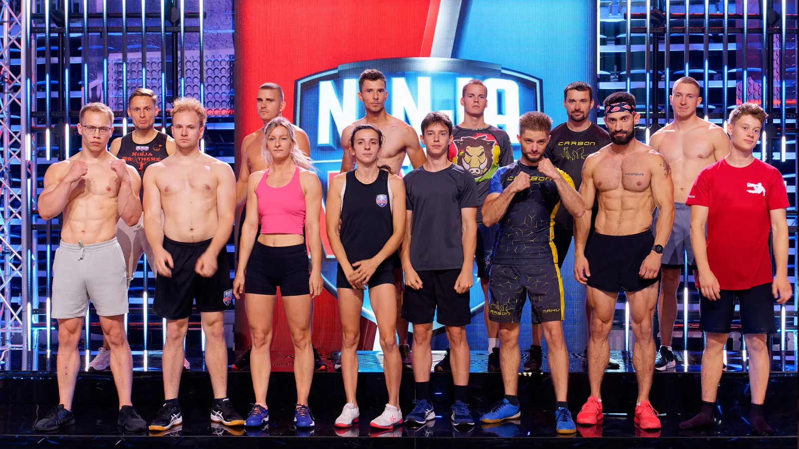 Grupa kilkunastu zawodników i zawodniczek 2. odcinka programu Ninja vs Ninja stojąca na scenie telewizyjnej przed startem. Uczestnicy w strojach sportowych gotowi do rywalizacji na ekstremalnym torze przeszkód.
