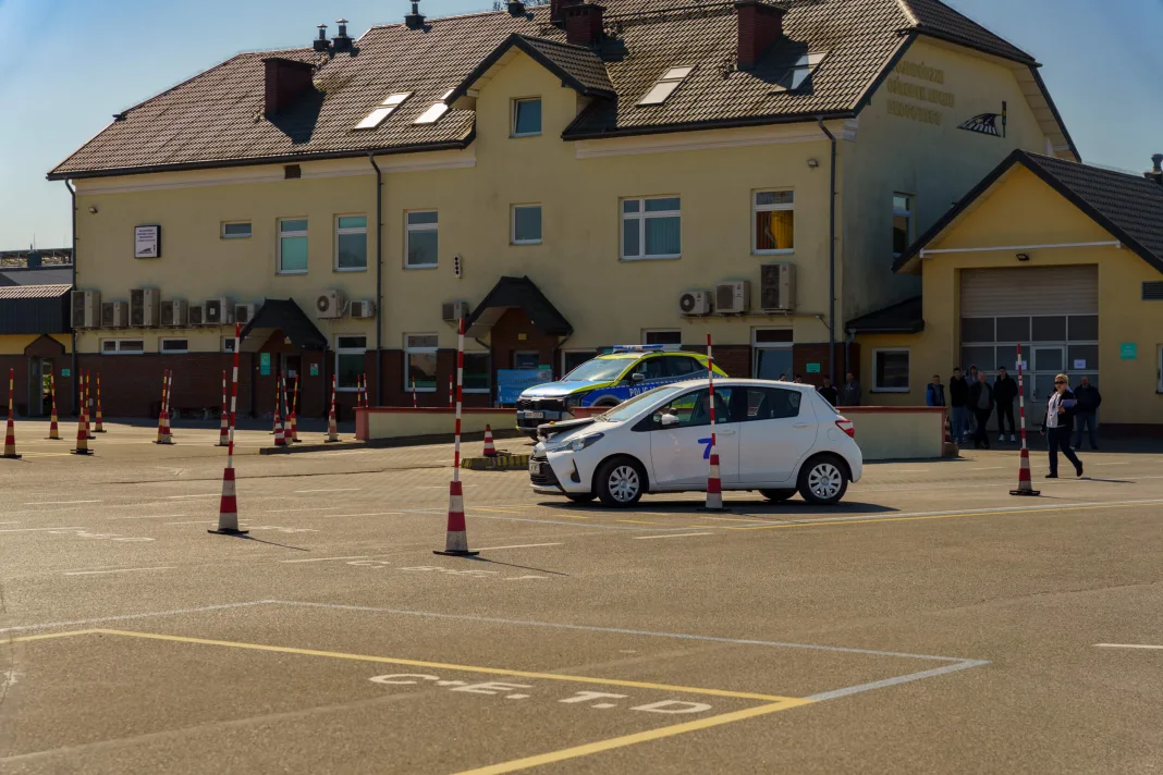 Fotografia placu manewrowego WORD w Polsce, na pierwszym planie biała Toyota Yaris z numerem '7' zaparkowana między pachołkami, w tle radiowóz policyjny i budynek WORD.